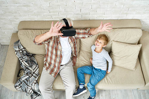 Portrait of excited senior man using VR glasses sitting on sofa at home with laughing grandson beside him