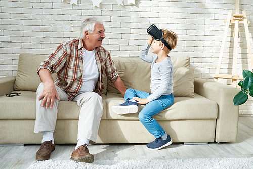 Portrait of happy senior man sitting on sofa having fun with little boy wearing VR glasses