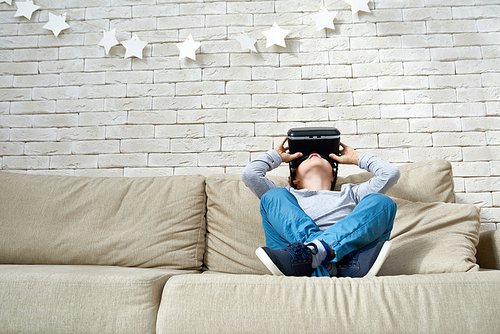 Low angle portrait of little boy wearing VR glasses watching 360 videos and playing augmented reality games at home sitting cross legged on cozy sofa, copy space