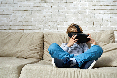 Full length portrait of little boy wearing VR glasses sitting cross legged on sofa, copy space