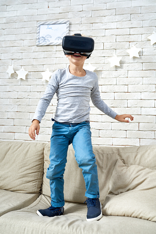 Portrait of excited little boy standing on sofa at home  wearing VR glasses watching 360 videos and playing augmented reality games
