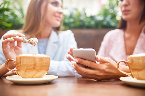 Portrait of two young women enjoying coffee at cafe table and chatting, focus on elegant female hand holding smartphone