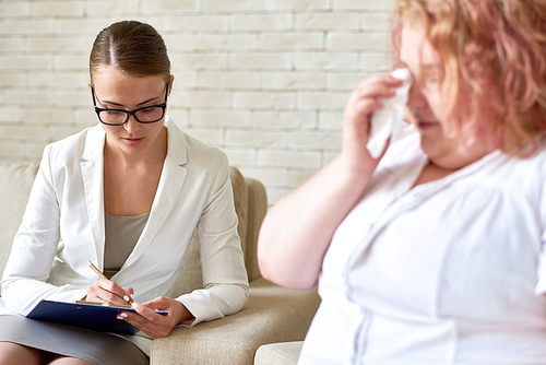 Portrait of beautiful young woman writing on clipboard while listening to  crying obese woman during therapy session on mental issues