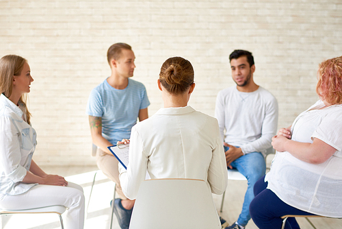 Back view of female coach working with group of young people sitting in circle during therapy session or training seminar in light spacious room