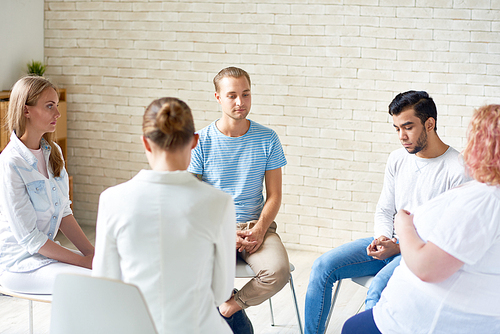 Back view of female coach working with group of young people sitting in circle during therapy session