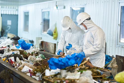 Portrait of two workers  wearing biohazard suits and hardhats working at waste processing plant sorting recyclable materials on conveyor belt