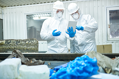Portrait of two workers  wearing biohazard suits working at waste processing plant using digital tablet and sorting trash standing by conveyor belt, copy space