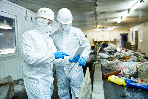 Portrait of two workers  wearing biohazard suits  using digital tablet  standing by conveyor belt at waste processing plant , copy space