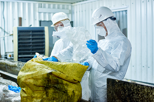 Portrait of two workers  wearing biohazard suits working at waste processing plant sorting recyclable plastic on conveyor belt