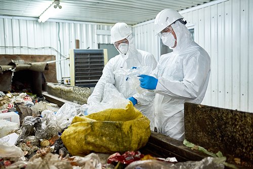 Portrait of two workers wearing biohazard suits sorting recyclable plastic and cardboard on conveyor belt at waste processing plant