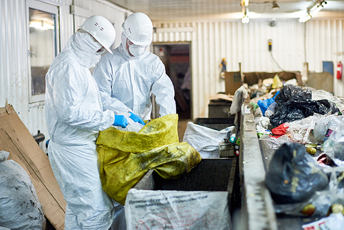 Side view  portrait of two workers wearing biohazard suits sorting recyclable plastic and cardboard on conveyor belt at waste processing plant