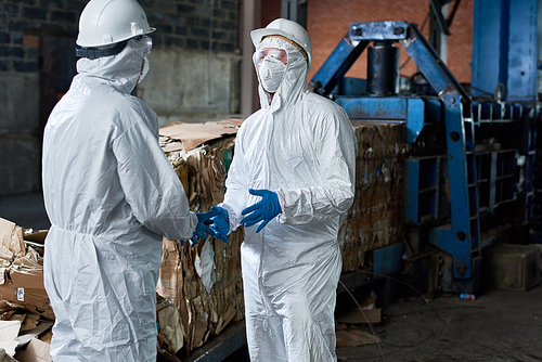 Portrait of two workers wearing biohazard suits communicating  in industrial warehouse of modern waste processing plant against recyclable cardboard in background, copy space