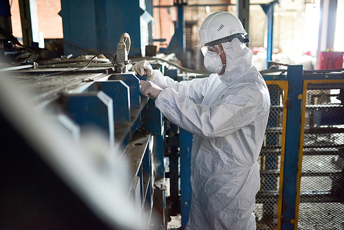 Side view portrait of worker wearing biohazard suit and hardhat standing in open workshop of industrial plant