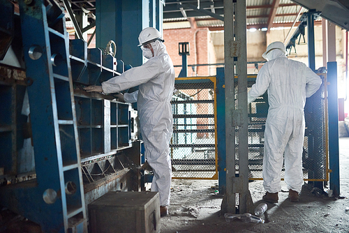Full length view of two workers wearing biohazard suit and hardhats working in open workshop of industrial plant