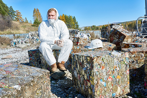 Portrait of young man wearing hazmat suit sitting on trash blocks outside waste processing factory, copy space