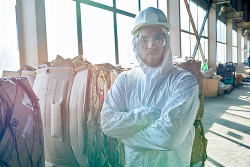 Portrait of young workers wearing biohazard suit standing in industrial warehouse of modern waste processing plant in sunlight, copy space