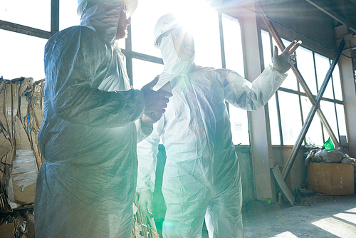 Portrait of two workers wearing biohazard suits standing in industrial warehouse of modern waste processing plant against window in sunlight