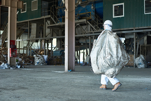 Full length portrait of man in hazmat suit carrying bag of recyclable materials in warehouse of waste processing plant, copy space
