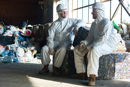 Full length portrait of two workers wearing hazmat suits sitting on blocks of packed trash in empty workshop of waste processing plant, copy space