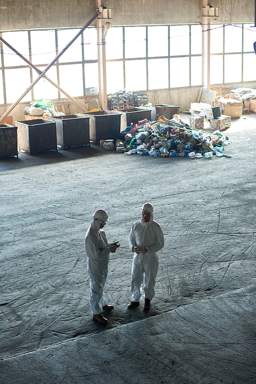 High angle view at two workers wearing warehouse suits standing in empty workshop of waste processing plant, piles of trash in background, copy space