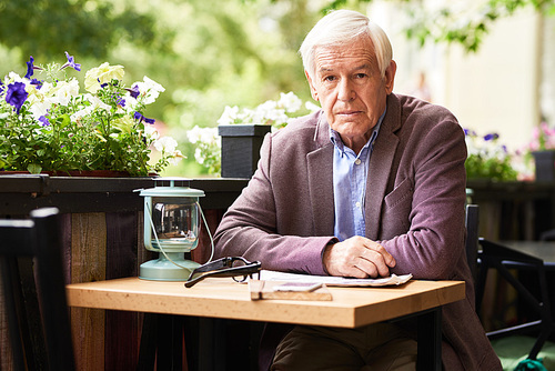 Portrait of thoughtful senior man sitting at table in outdoor cafe lounge with newspaper, 