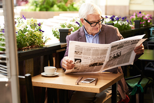 Portrait of senior man wearing glasses reading morning newspaper in cafe outdoors