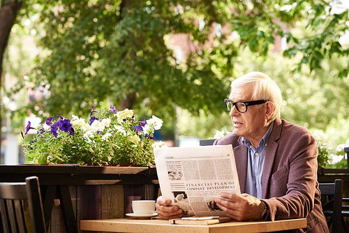 Portrait of senior man wearing glasses reading morning news in cafe outdoors enjoying sunny day in retirement
