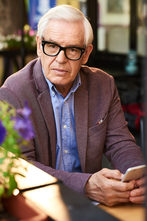 Portrait of modern senior man using smartphone in outdoor cafe, looking away thoughtfully