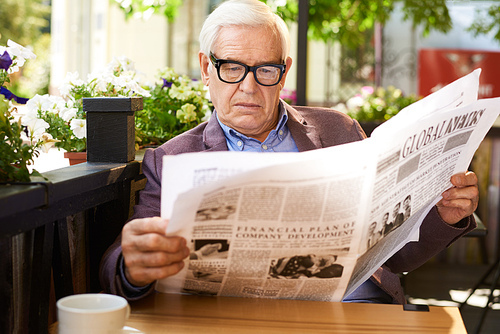 Portrait of modern senior man wearing glasses reading newspaper in cafe outdoors