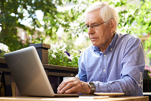 Portrait of senior man working with laptop in outdoor cafe lounge, looking at computer screen
