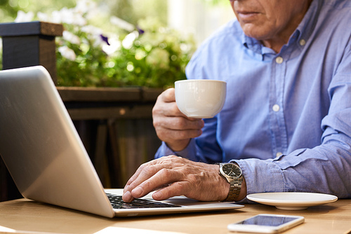 Closeup image of unrecognizable senior man using laptop computer in cafe outdoors holding coffee cup