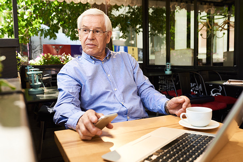 Portrait of senior man working with laptop in outdoor cafe lounge, looking away pensively