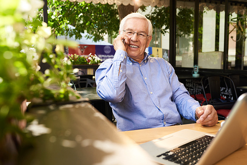 Portrait of senior man speaking by phone smiling happily while working with laptop in outdoor cafe lounge during coffee break