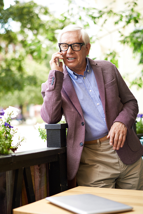 Portrait of modern senior man speaking by phone outdoors in cafe, leaning on flowerbed