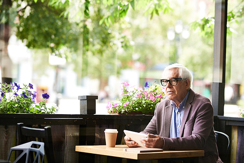 Portrait of pensive senior man sitting at cafe table outdoors holding digital tablet, copy space