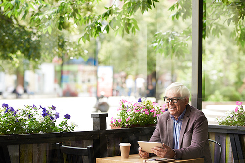 Portrait of smiling senior man sitting at cafe table outdoors holding digital tablet, copy space