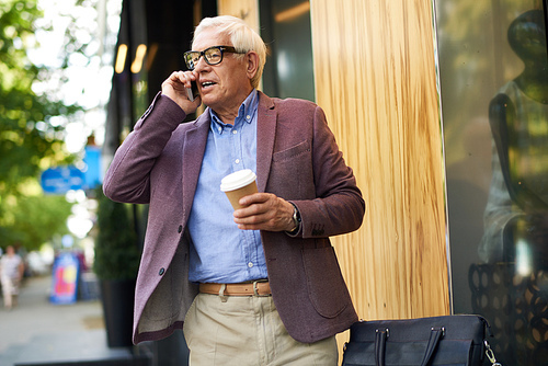 Portrait of modern senior man using smartphone outdoors, in city street holding coffee cup in one hand