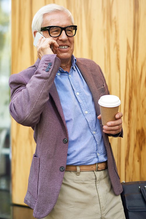 Portrait of modern senior man speaking by phone and smiling outdoors in city holding coffee cup in one hand