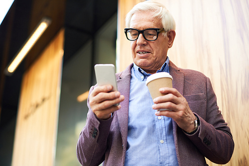 Portrait of modern senior man using smartphone outdoors, in city street holding coffee cup in one hand