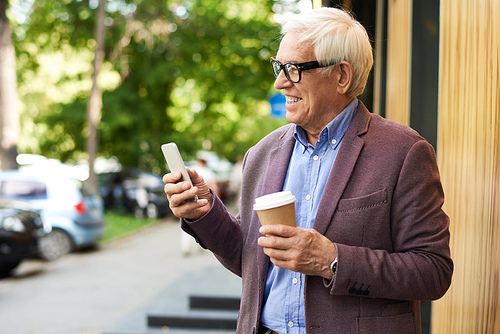 Portrait of modern senior man using smartphone outdoors and smiling, standing in city street holding coffee cup in one hand