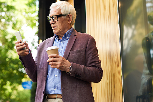 Portrait of modern senior man using smartphone outdoors in city, holding coffee cup