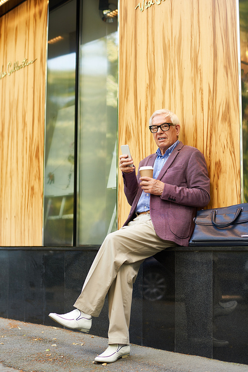 Portrait of trendy senior man relaxing outdoors with cup of coffee and modern smartphone, 