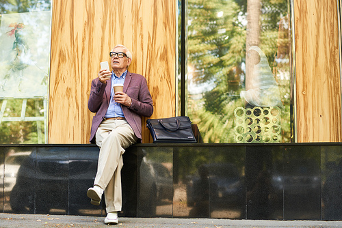 Portrait of trendy senior man relaxing outdoors with cup of coffee and modern smartphone