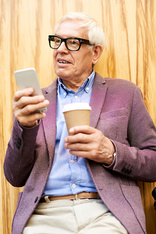 Portrait of trendy senior man relaxing outdoors holding smartphone and cup of coffee