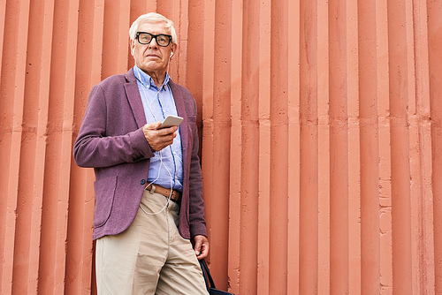 Portrait of trendy senior man holding smartphone posing outdoors standing by bright orange wall