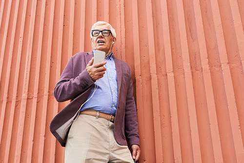 Low angle portrait of trendy senior man using smartphone outdoors standing by bright orange wall