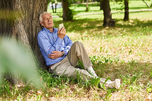 Portrait of modern senior man relaxing listening to music from smartphone  in park under tree