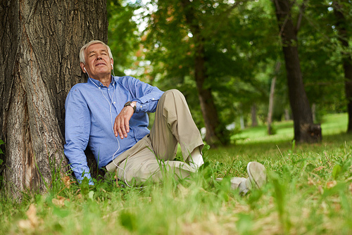 Portrait of modern senior man relaxing listening to music from smartphone  in park sitting on grass with eyes closed