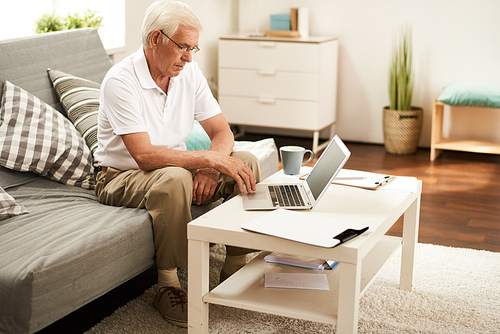 Portrait of modern senior man using laptop at home, working in retirement