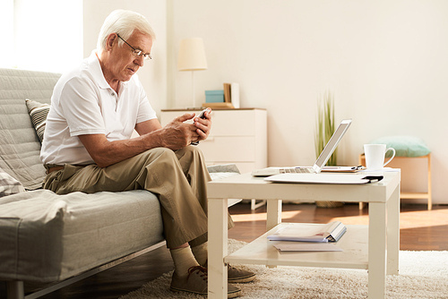Portrait of modern senior man learning to use smartphone and laptop at home in cozy living room